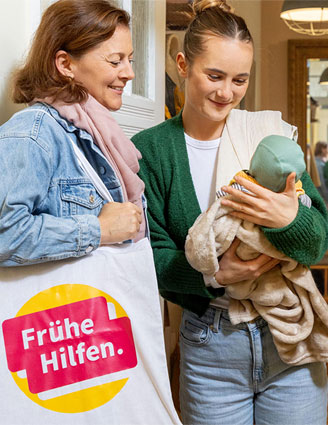 Junge Mutter begrüßt Besuchende der Frühen Hilfen an der Haustür und betrachtet mit ihr neutral das Baby