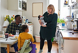 Familie beim Frühstück; Mutter steht mit Baby und schaut zu Vater und Kleinkind, die am Tisch sitzen