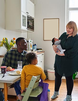 Familie beim Frühstück; Mutter steht mit Baby und schaut zu Vater und Kleinkind, die am Tisch sitzen