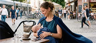 Stillende Frau sitzt mit Baby im Außencafé und trägt einen blauen Umhang